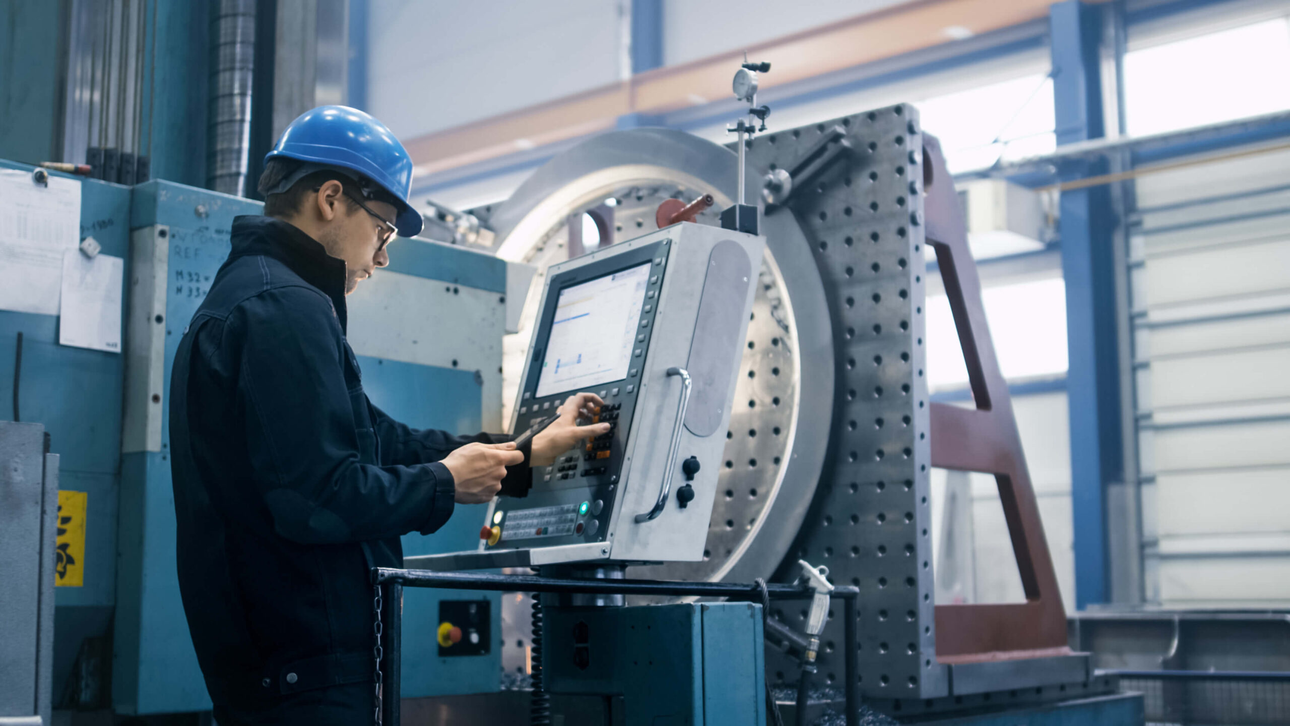  A male operator wearing a hard hat uses a control panel to operate heavy machinery in a manufacturing facility.